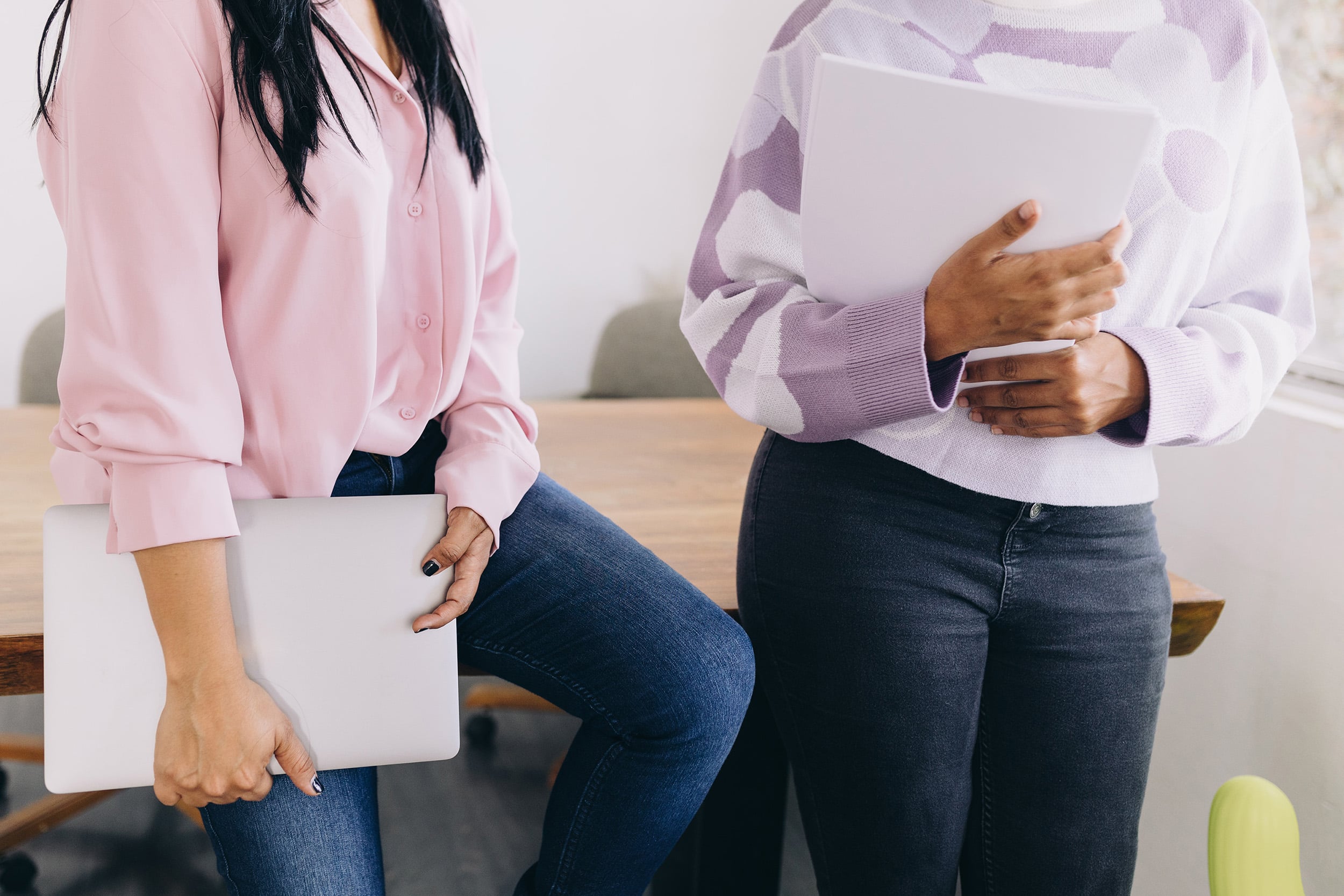 Two women standing together in a workspace; one is holding a laptop. The image is cropped to show only their torsos