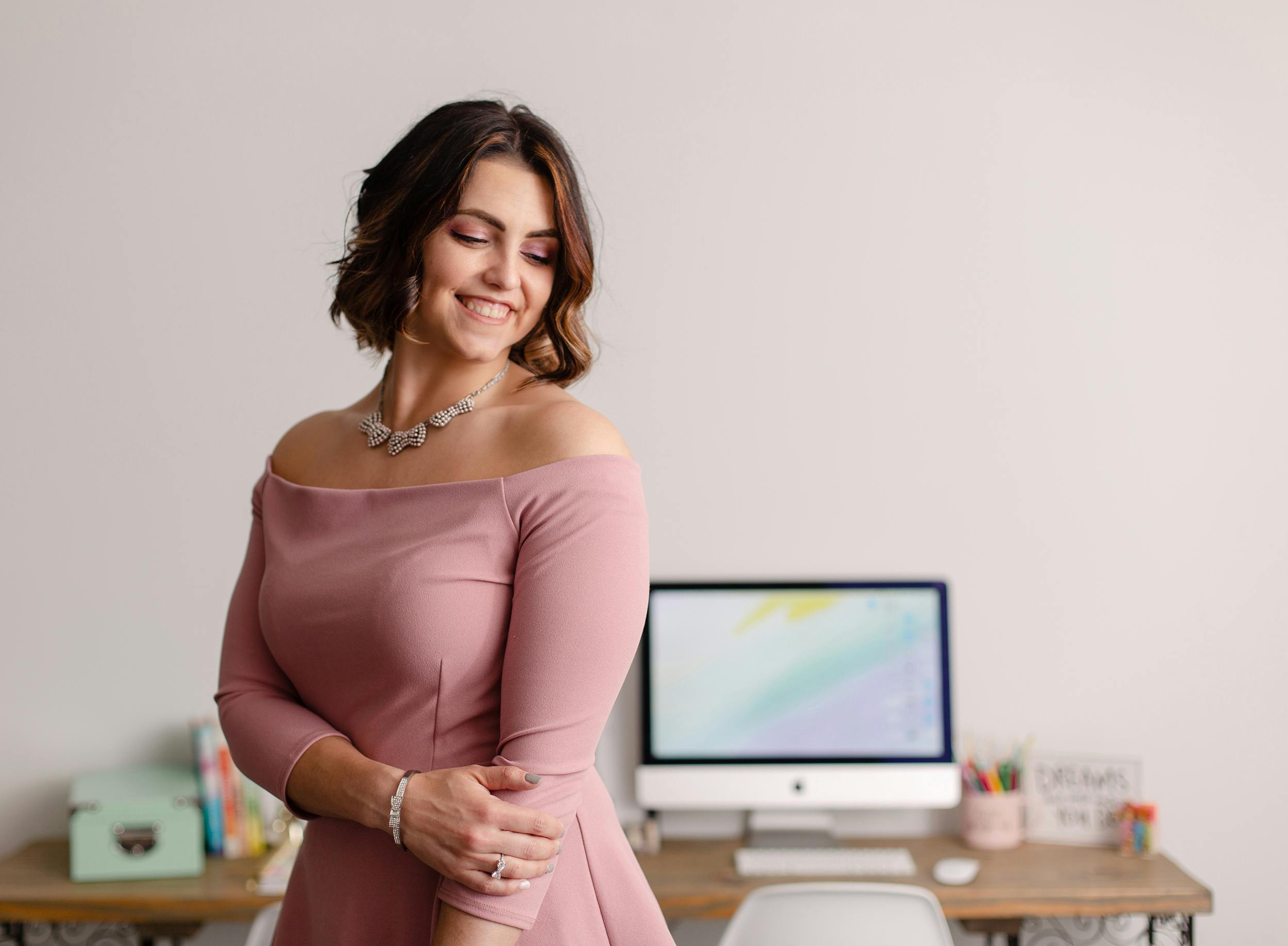 Megan in a pink shirt standing in front of an iMac on a desk, looking down and smiling